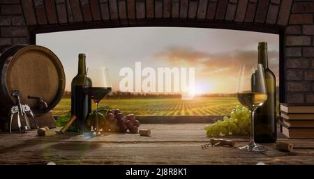 Still life with wine glasses, bottles, grapes on wooden table in wine cellar. Panoramic window view of lush vineyards at sunset. Stock Photo