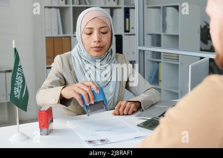 Young adult Muslim consular officer working in Saudi Arabia embassy giving approval for travel visa to Caucasian man Stock Photo
