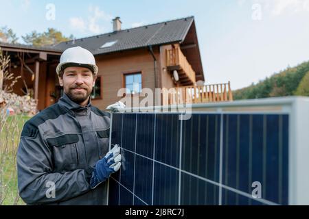 Smiling handyman solar installer carrying solar module while installing solar panel system on house. Stock Photo