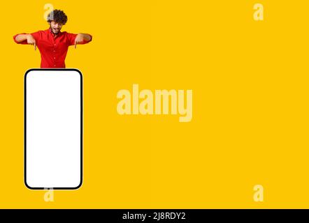 Excited curly Indian young man pops up like gin above smartphone and points finger down at empty screen isolated on yellow background, latin guy presenting and advertising mobile app, copy space Stock Photo