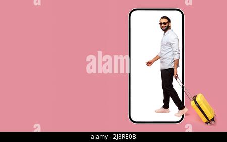 Great mobile app for tourism and travel. Cheerful carefree Indian man in sunglasses walking with luggage into huge smart phone isolated on pink, advertising of booking platform, copy space, mock up Stock Photo