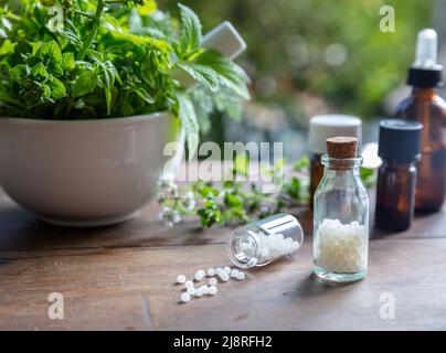 Homeopathy pharmacy, herbal, natural medicine. Homeopathic globules scattered out of a glass bottle, fresh herb in a mortar, green nature background. Stock Photo