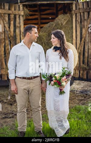 Stylishly dressed handsome groom with a beard in black sunglasses hugs wife. Bride in a dress with a bouquet puts a hat on and laughs. Rustic wedding Stock Photo
