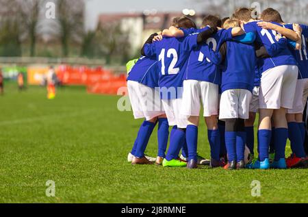 Kids soccer team with coach in group huddle before the match. Elementary age children are listening together to coach motivational speech. Boys in blu Stock Photo