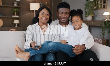 Carefree african american father, mather relaxing with biracial daughter child girl at hme holidays sitting on sofa at home watching television togeth Stock Photo
