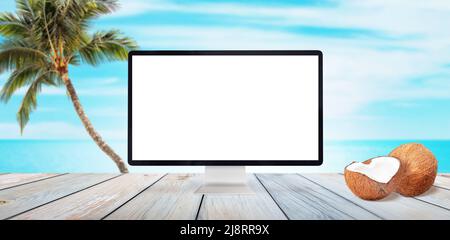 Computer display mockup on wooden table. Beach, palm and sea in background. Coconuts beside. Isolated white display for summer travel promotion Stock Photo