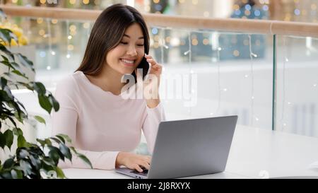 Happy Asian Korean woman freelancer student businesswoman holding mobile phone call working with laptop multitasking girl making online order communic Stock Photo
