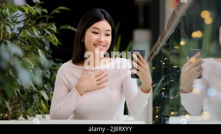 Asian korean friendle girl japanese lady chinese female smiling waving hello to phone webcam woman with mobile gadget talking make video call in cafe