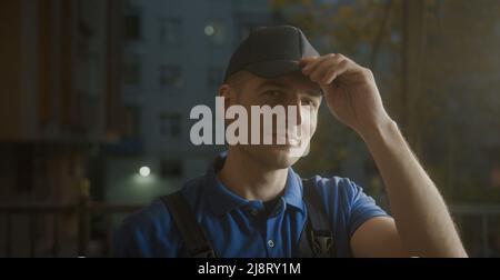 Attractive young delivery man courier in wearing the blue hat on his head put your hands together and smiling and talking into camera Stock Photo