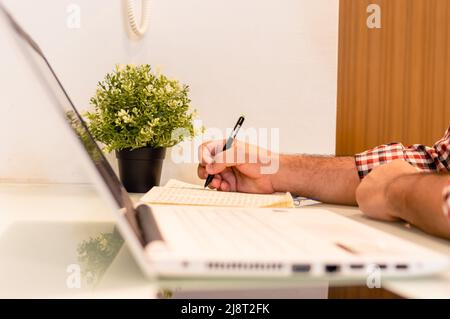 side view unrecognizable man working at home with the laptop on the table and writing down data in a notebook with a pen. copy space. Stock Photo