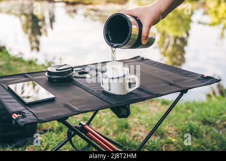 Pouring hot water through trendy convenient paper drip coffee bag into metal cup on camping table outdoors. Making freshly brewed coffee in nature Stock Photo