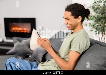 Young handsome Indian freelancer man using smartphone for online chatting in social media, using mobile app, shopping or texting online. Happy guy using phone, scrolling news feed sitting indoor Stock Photo