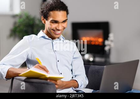 Happy young curly man using laptop computer for studying and work on the distance. Guy watching webinar, taking educational course online, while sitting on the couch at home Stock Photo