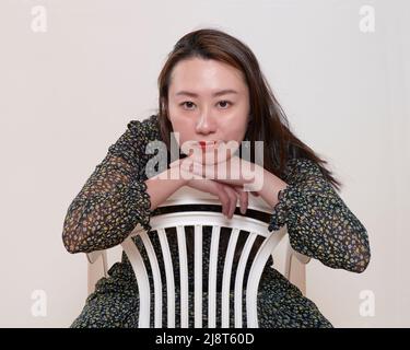 Young Hong Kong Chinese woman wearing long-sleeved patterned dress and sitting reversed way round on white chair in front of pale grey backgound Stock Photo