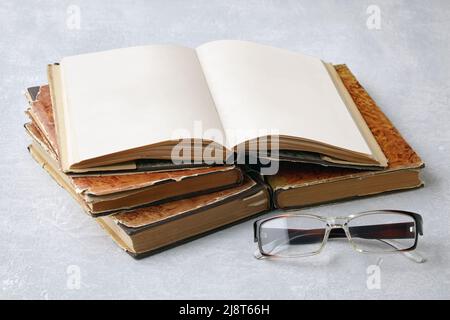 Open vintage book notebook with blank pages and glasses on a stack of old shabby books on a gray concrete table Stock Photo