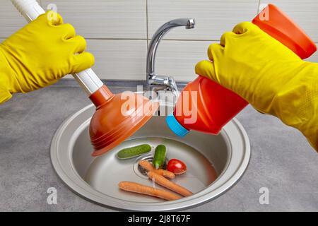 Hands in yellow rubber gloves hold a plunger and a pipe cleaner against the background of a clogged kitchen sink with vegetables cucumbers, tomatoes, Stock Photo