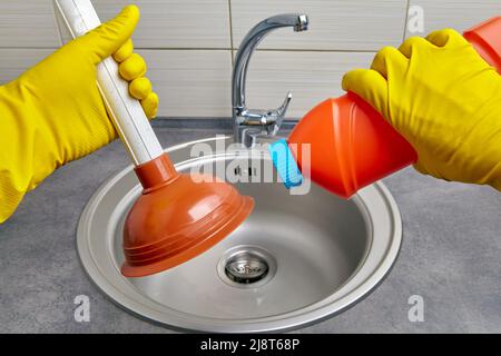 Hands in yellow rubber gloves hold a plunger and a pipe cleaner against the background of a kitchen sink. Housework cleaning a clogged sewer drain Stock Photo