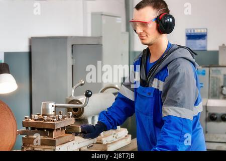 Young turner in safety glasses and headphones works at lathe in workshop. Authentic scene workflow. Caucasian worker processes metal parts. Stock Photo