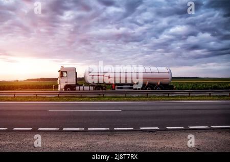 A truck with a semi-trailer tanker transports dangerous goods against the backdrop of a sunny sunset and the sky in the clouds. Transportation of flui Stock Photo