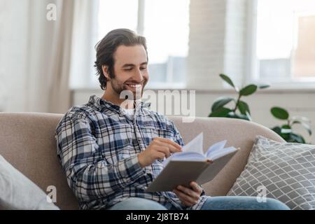 Happy millennial handsome caucasian guy with stubble sits on sofa reads book in living room interior Stock Photo