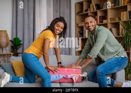 Happy black couple preparing for their trip, packing things together and trying to close suitcase at home