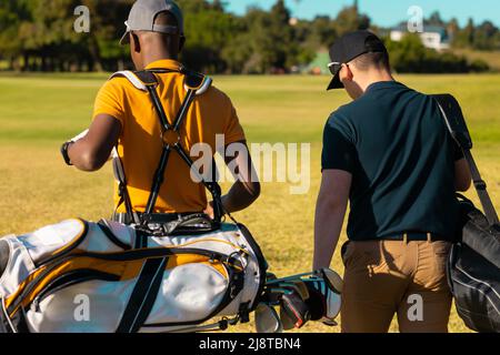 Rear view of multiracial young male friends wearing caps with golf bags walking at golf course Stock Photo