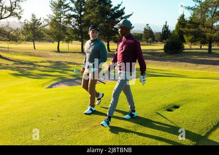 Multiracial young male friends with golf clubs talking while walking at golf course in summer Stock Photo