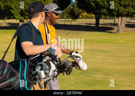 Side view of multiracial young male friends wearing caps with golf bags walking at golf course Stock Photo