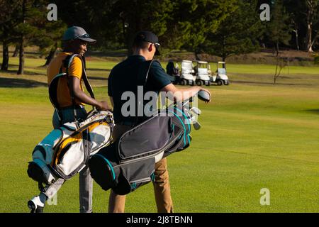 Young male multiracial friends wearing caps with golf bags walking at golf course in summer Stock Photo