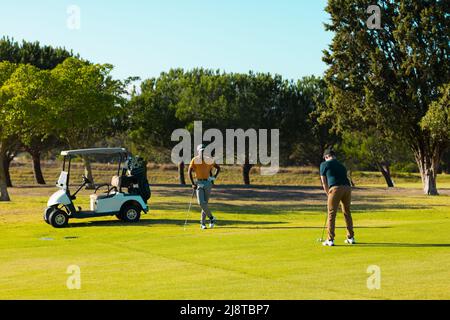 Multiracial young male friends playing golf against trees at golf course on sunny day, copy space Stock Photo