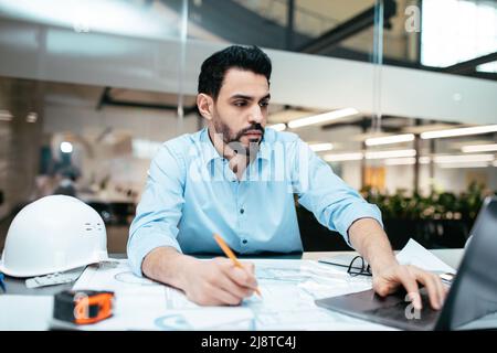 Concentrated busy handsome millennial middle eastern man architect with beard working on project with laptop Stock Photo