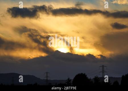 Evening sun setting behind dark clouds over silhouetted electricity pylons, UK. Stock Photo