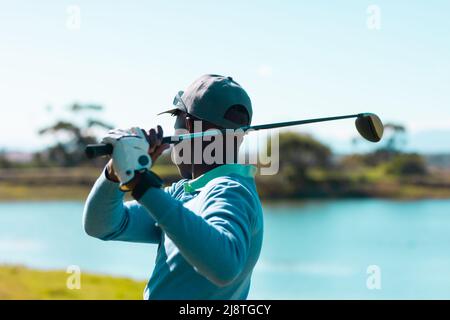 Side view of african american young man taking golf shot with club against lake and clear sky Stock Photo