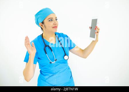 beautiful woman doctor, wearing blue uniform and blue surgical cap, stethoscope on her neck, waving at the tablet on white background Stock Photo