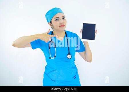beautiful woman doctor, wearing uniform and blue surgical cap, stethoscope on her neck, pointing at the tablet on white background Stock Photo