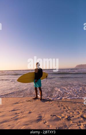 African american bearded senior man with surfboard standing against sea and clear blue sky Stock Photo