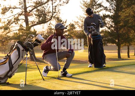 Multiracial young male friends with golf clubs and bags at golf course against trees during sunset Stock Photo