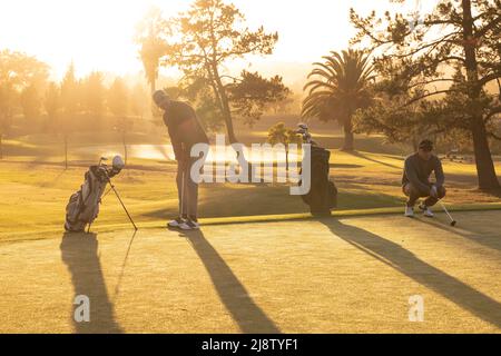 Multiracial young male friends playing golf on grassy land against trees and sky at golf course Stock Photo