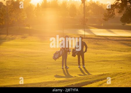 Full length of multiracial young male friends with golf bags standing at golf course during sunset Stock Photo