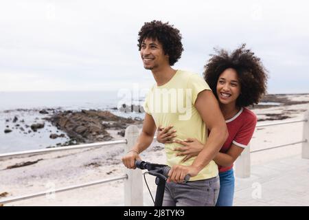 Young african american woman enjoying push scooter ride with afro boyfriend by beach, copy space Stock Photo
