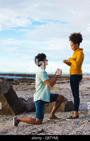 Young afro man with ring box proposing african american girlfriend while kneeling at beach Stock Photo