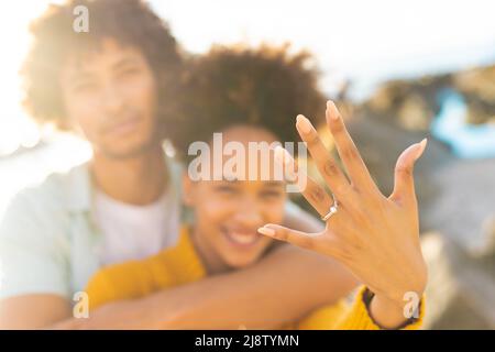 Cheerful excited woman showing off her engagement ring with african american boyfriend at beach Stock Photo