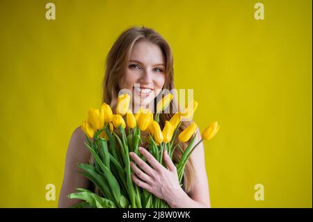 Caucasian woman with an armful of yellow tulips on a yellow background. International Women's Day. Bouquet of spring flowers Stock Photo