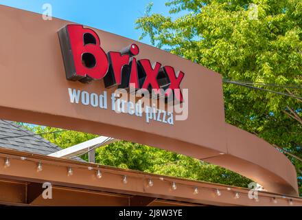 Exterior facade brand and logo signage for 'Brixx' wood fired pizza store on a bright sunny day with shadows, green trees leaves and a clear blue sky. Stock Photo