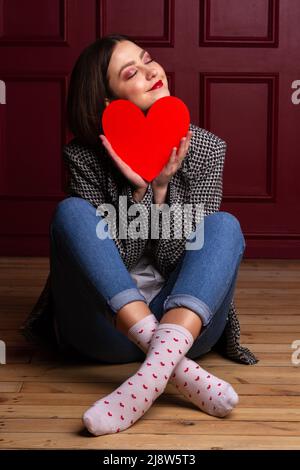 Dreamy smiling short-haired woman in jacket sitting on wooden floor legs crossed and red background holding red heart shape in front of her chin Stock Photo