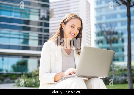 Carefree female office employee typing on the laptop sitting on the step in the city, businesswoman or freelancer woman responds to emails, working outdoors, urban landscape on the background Stock Photo