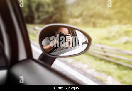man takes photo in mirror in car. A man is taking photo someone or something from an open car window. Reflection in the side mirror of the car. Stock Photo