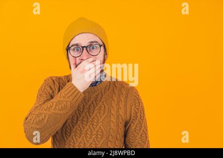 Studio shot on orange background of an isolated shocked scared surprised caucasian man looking at camera with his eyes wide open covering his mouth with his hand wearing a sweater and a hat. High quality photo Stock Photo