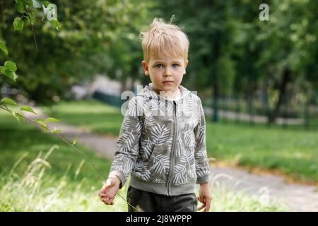 A portrait of a cute blond little boy in a green city park during the summer. Stock Photo