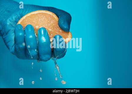 Close-up shot of fresh juice dripping from ripe half of orange while male hand squeezing it against blue background Stock Photo